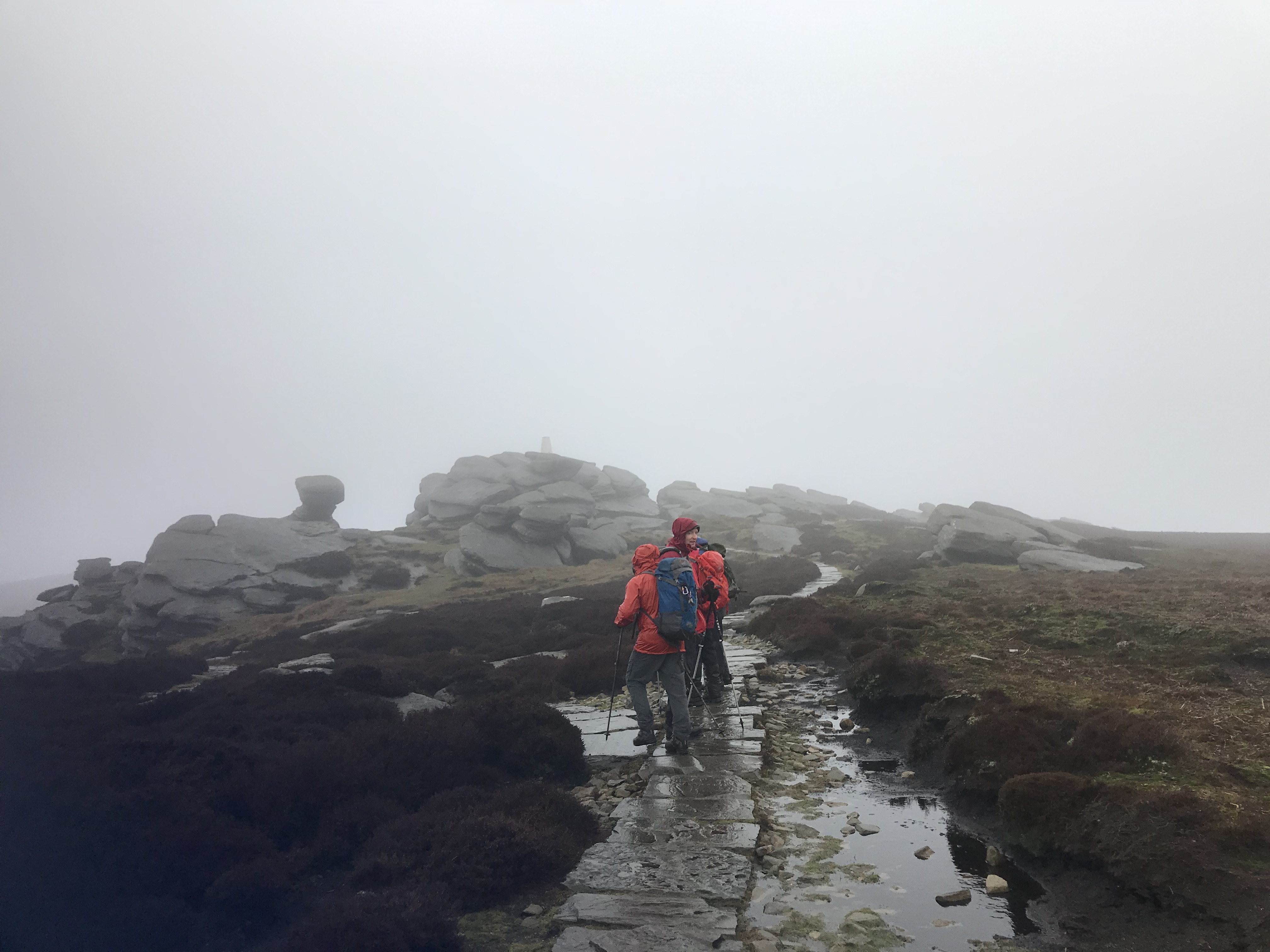 A group of walkers in wet weather gear stand on a rock path amid bog and heather.  Mist shrouds the rocky outcrop ahead.