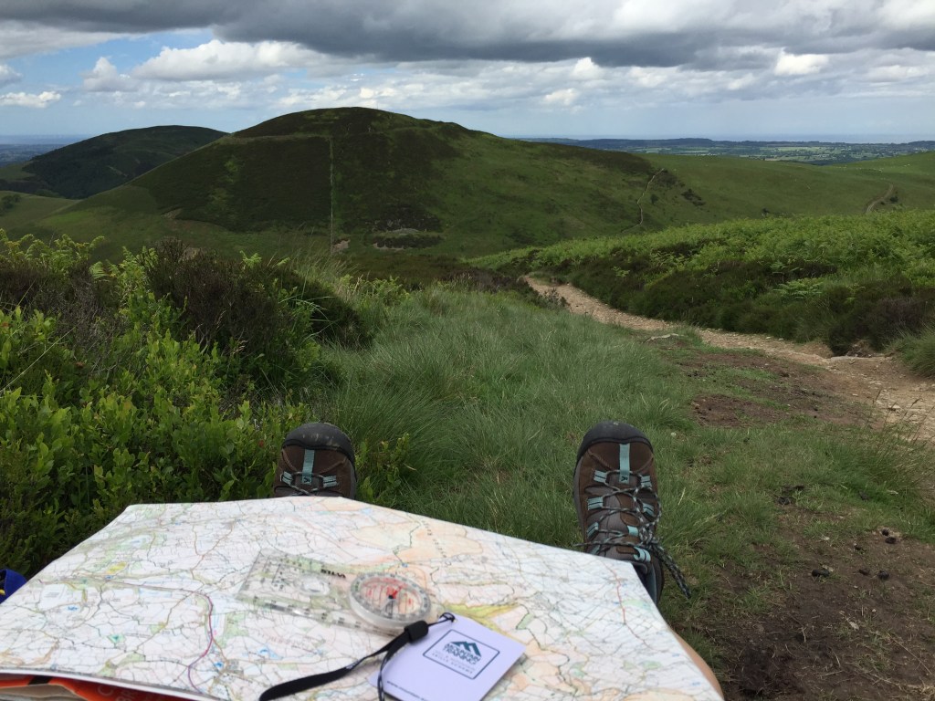 A map and compass on a walkers lap, looking towards a range of hills