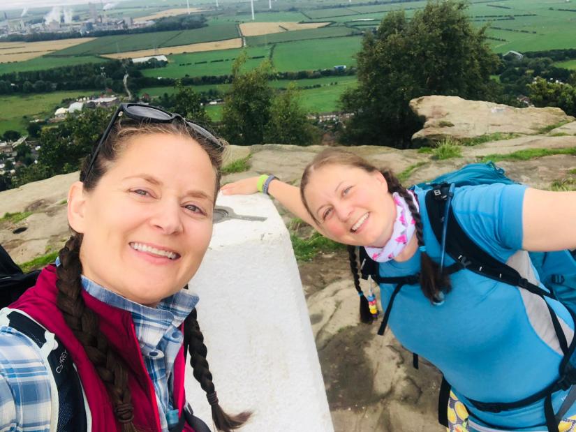 A selfie of two women in walking kit next to a trig on a hill