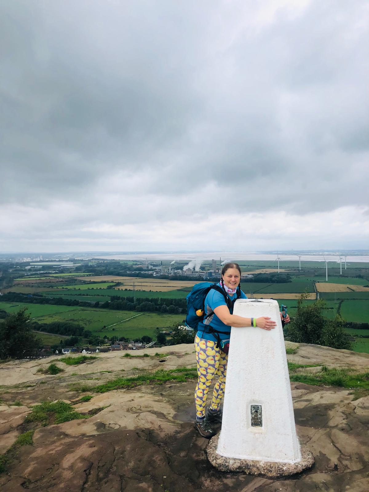 a woman in walking kit hugging a trig on a hill overlooking a windfarm