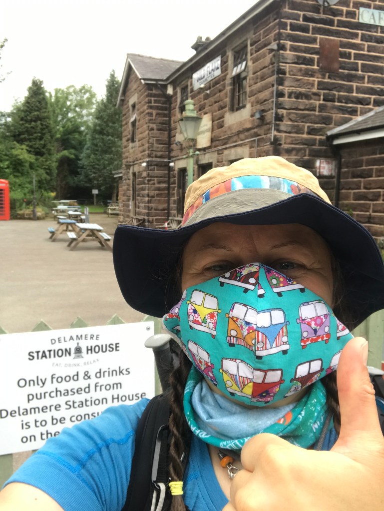 A selfie of a woman wearing a mask outside a stone building, giving a thumbs up sign