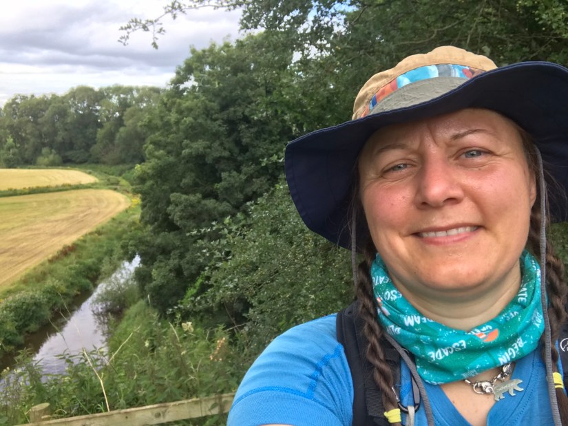 a selfie of a woman in outdoor gear with trees, a river and harvested fields in the background