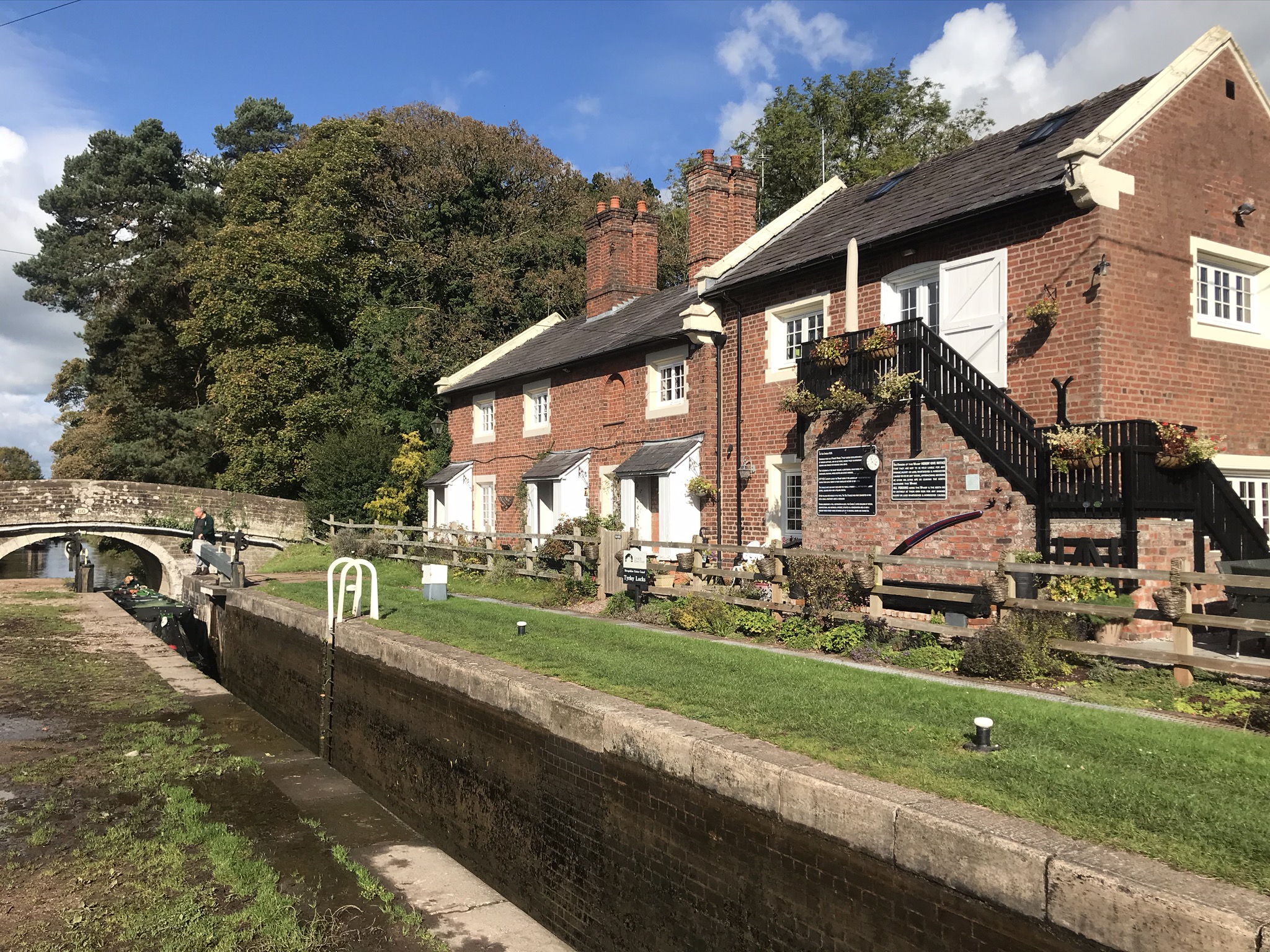 A two-story red brick building with hanging baskets of flowers and white painted windows over looking a canal lock. the sky is blue and sunny, a narrowboat is entering the bottom of the lock.