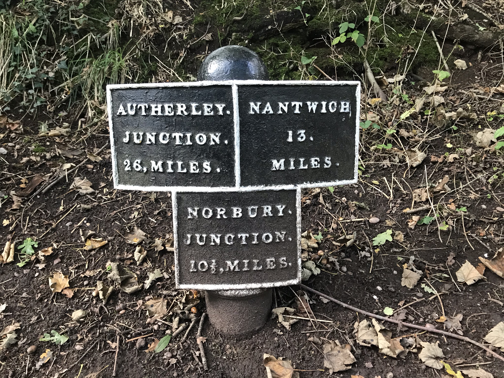 A black metal sign made of three squares with white writing in a soil verge. The top left sign is "Autherley Junction 26 miles", the top right is "Nantwich 13 miles" and the middle bottom is "Norbury junction 10 1/2 miles"