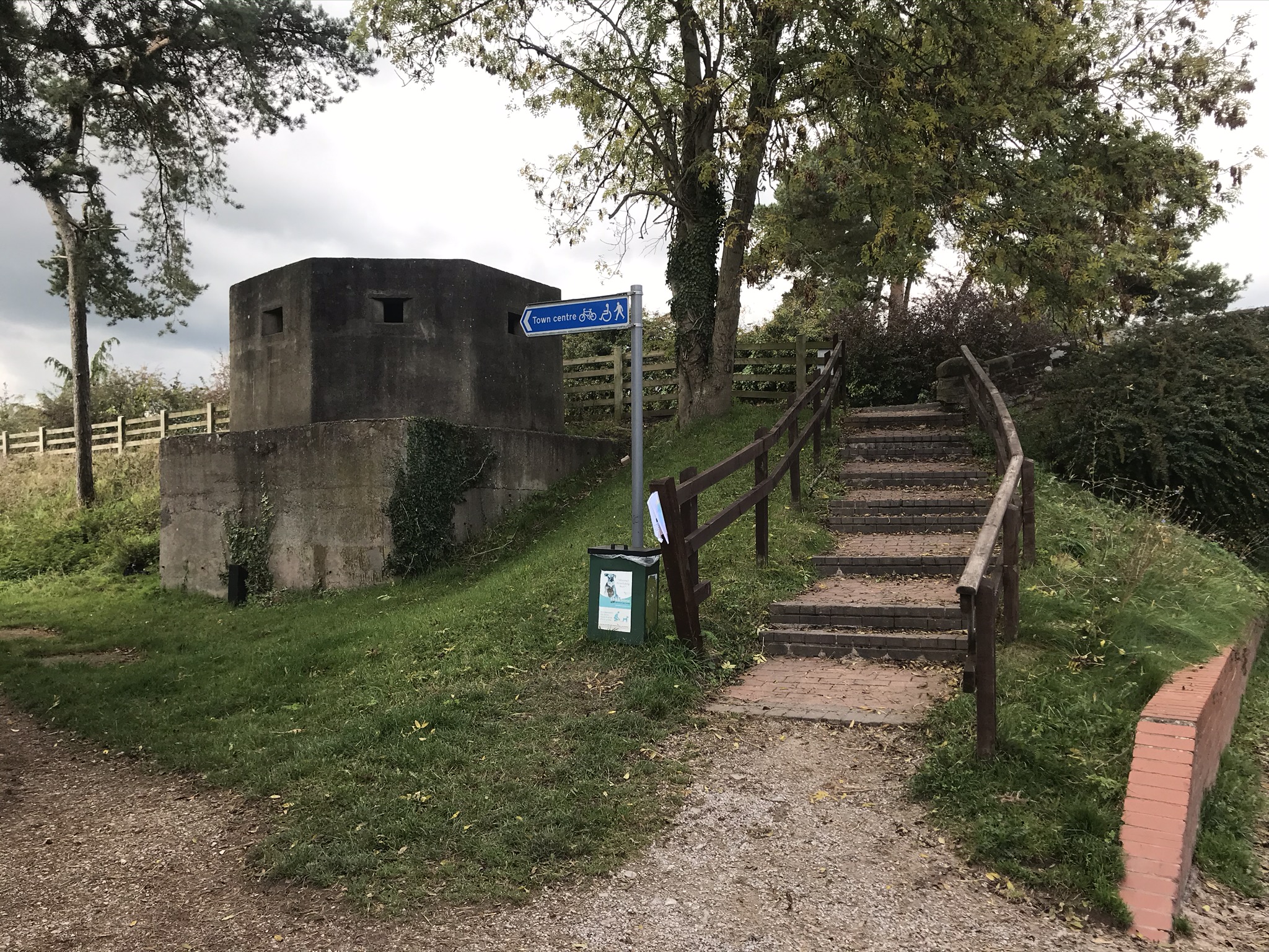 A concrete WW2 pillbox next to brick steps and a blue sign pointing towards the town centre