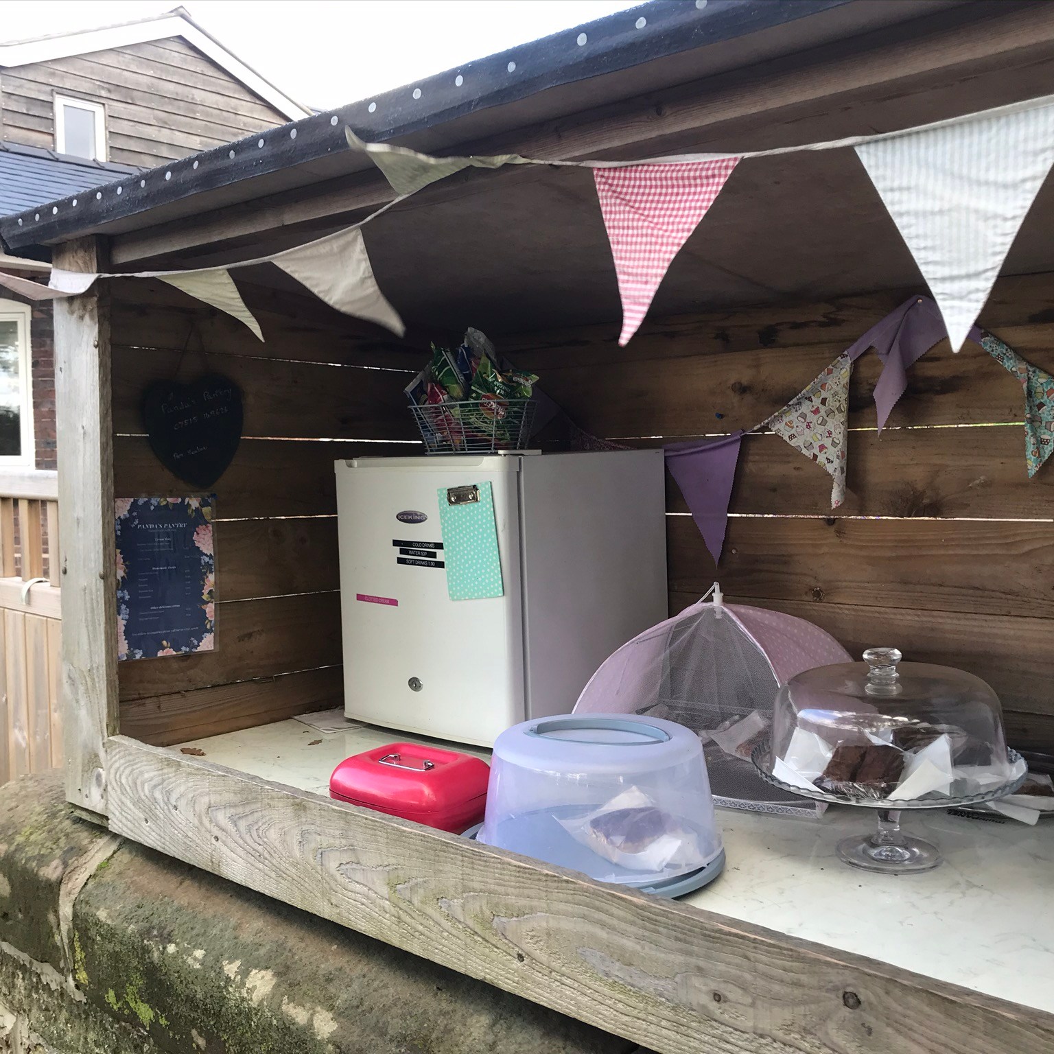 A timber shelter with pastel coloured bunting. There is cake under covers, a fridge and a red cash box.
