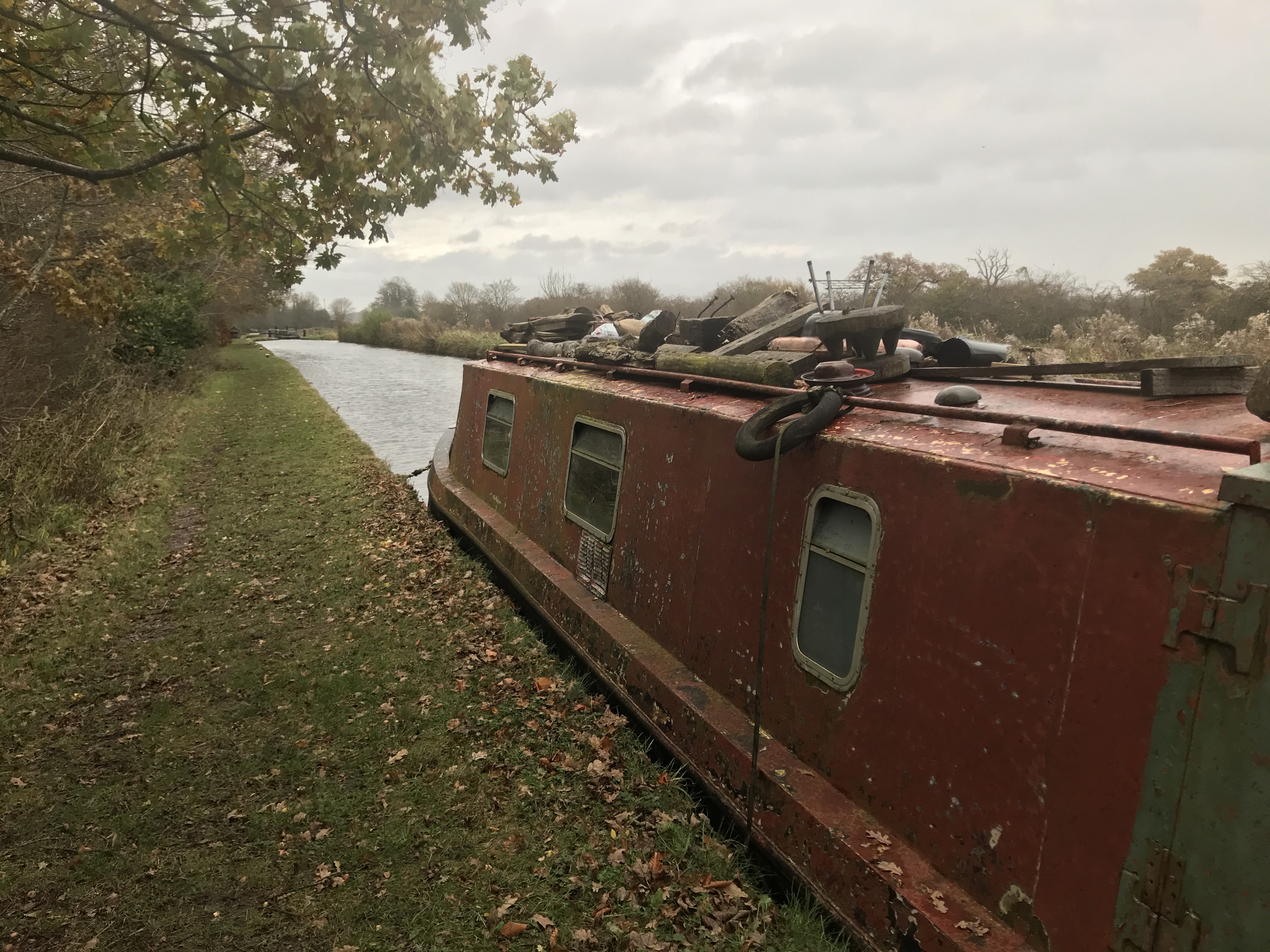 A rusty red narrowboat moored along a grassy towpath. The boat's windows are cracked open and there are logs, tyres and a BBQ on the roof. Leaves have fallen from the trees and the sky is overcast