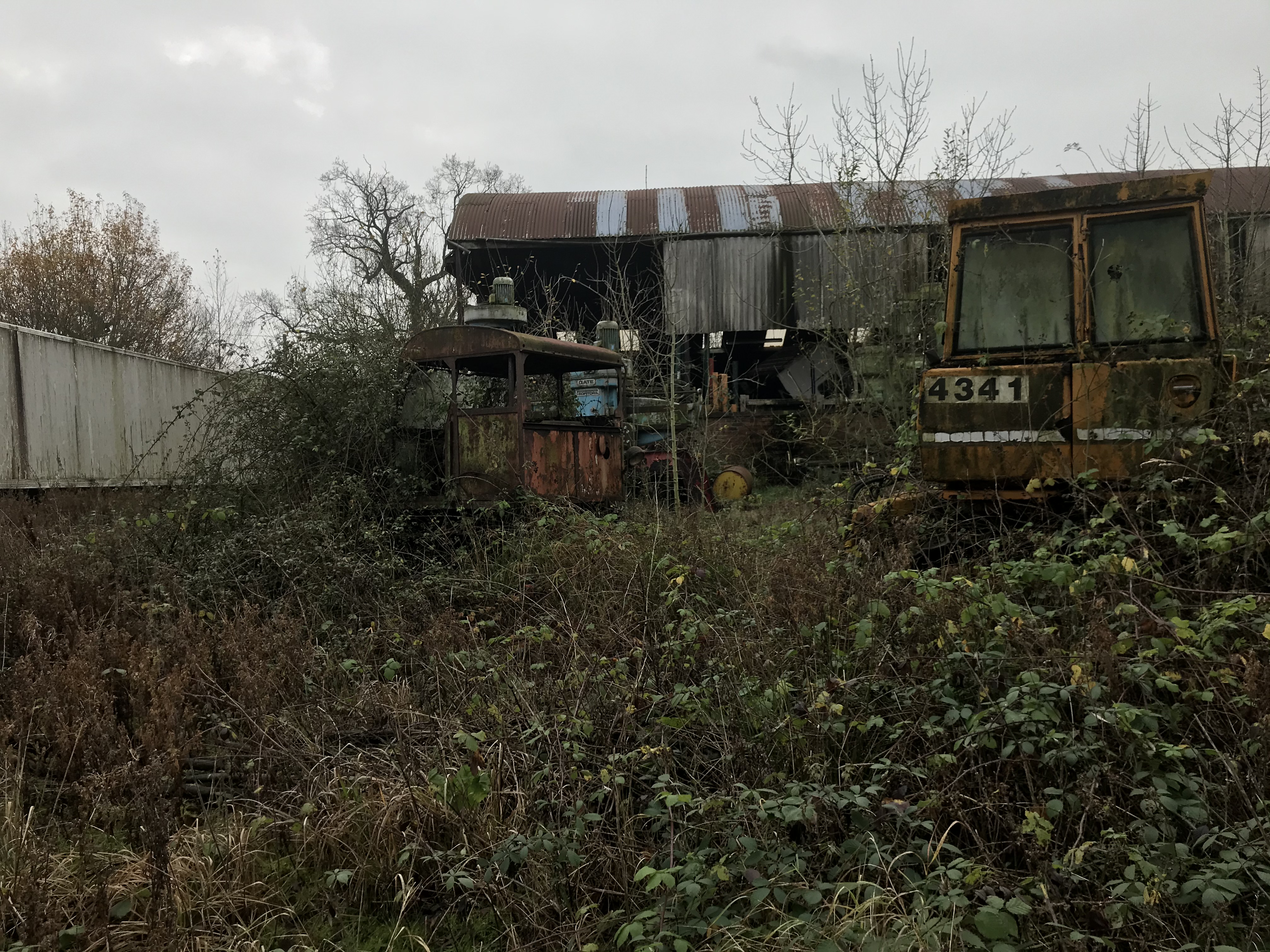A rusty corrugated steel barn with rusty agricultural and railway maintenance machinery, all heavily overgrown with thick brambles