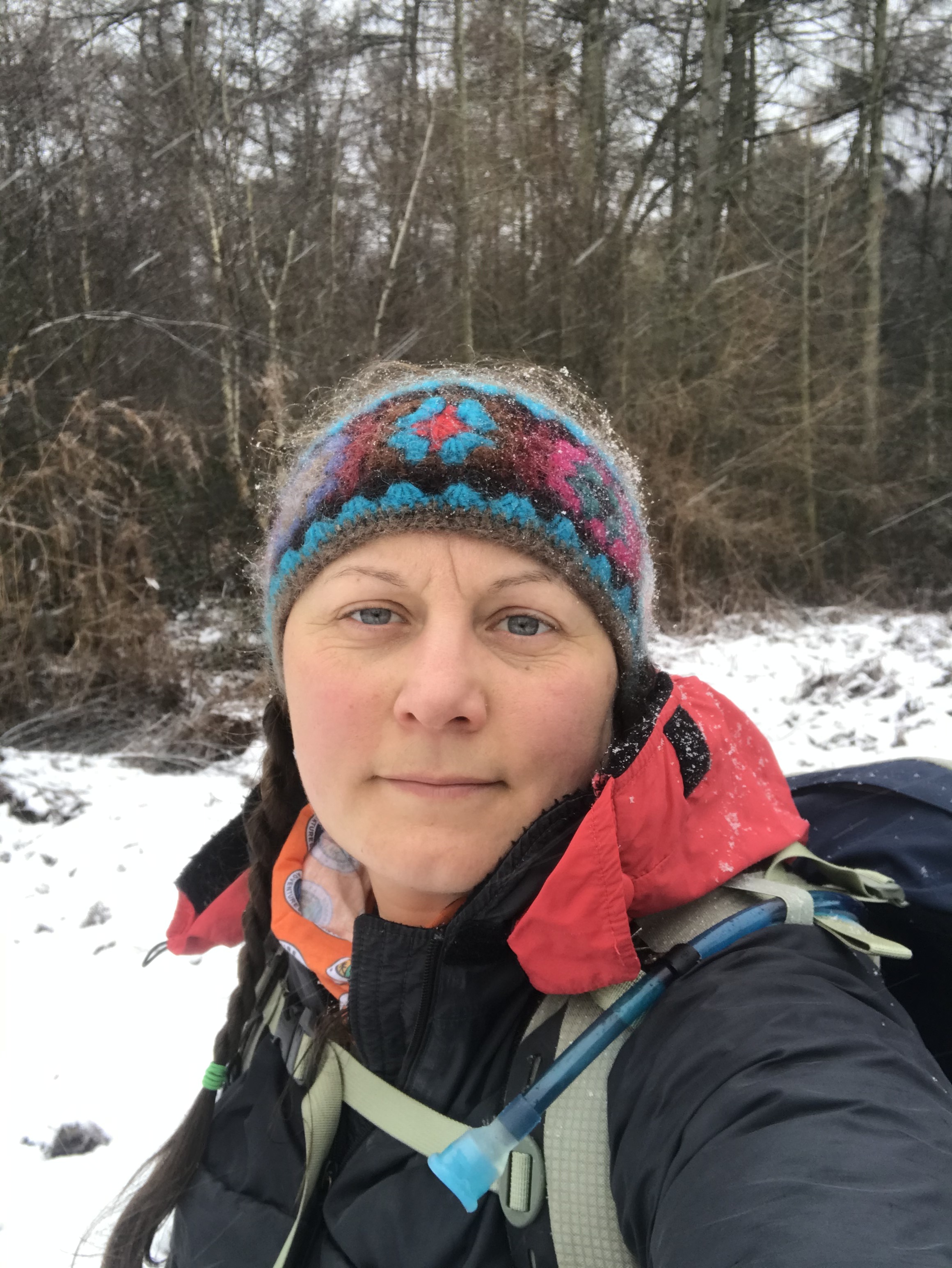 A selfie, wearing winter walking gear. The ground is covered in snow with wood in the background.