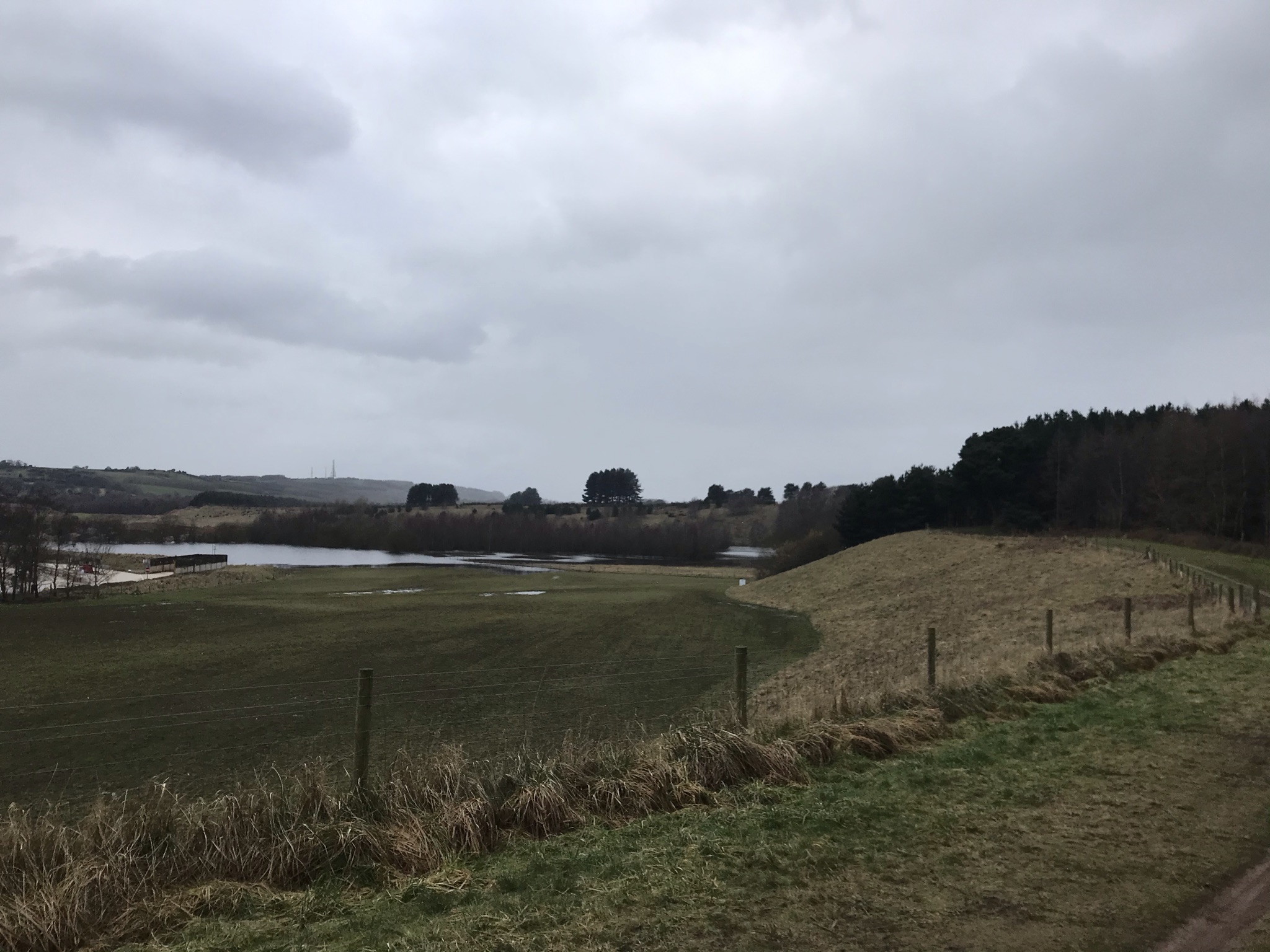 Grass banks with a lake in the distance. A fence run along the top of the bank with woods to the right. In the distance there are hills with a mast on top