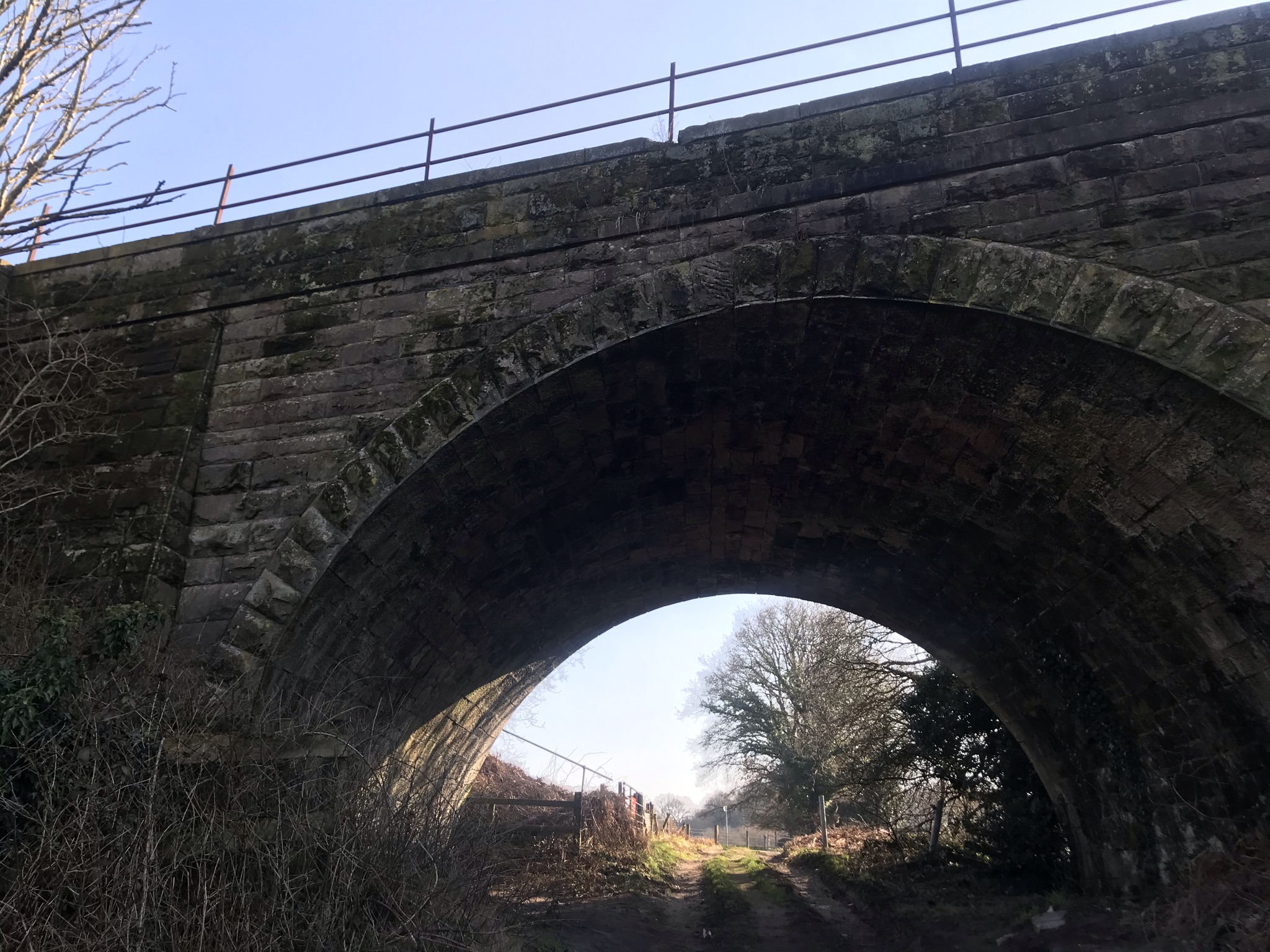 Looking up at a stone arch bridge. There is a rutted track underneath with bare trees in the background