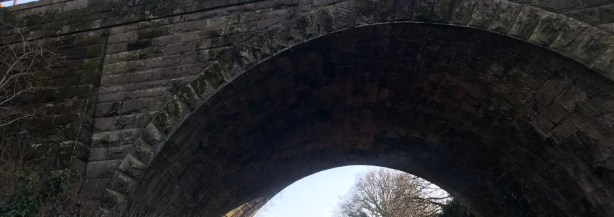 Looking up at a stone arch bridge. There is a rutted track underneath with bare trees in the background