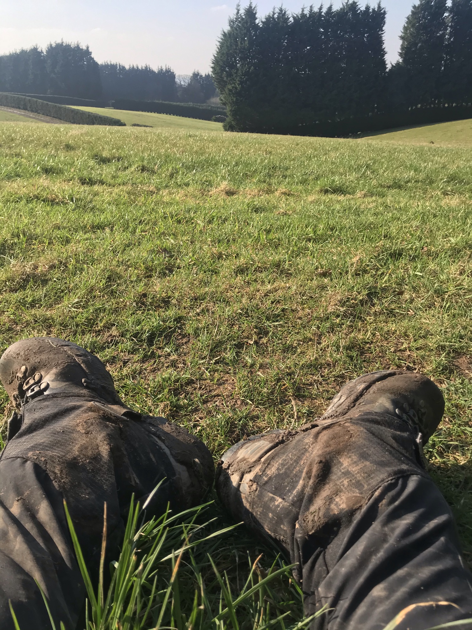 muddy boots of a person sat on the grass. The fields are gently rolling in the background and there are tall thick trees and neat hedges