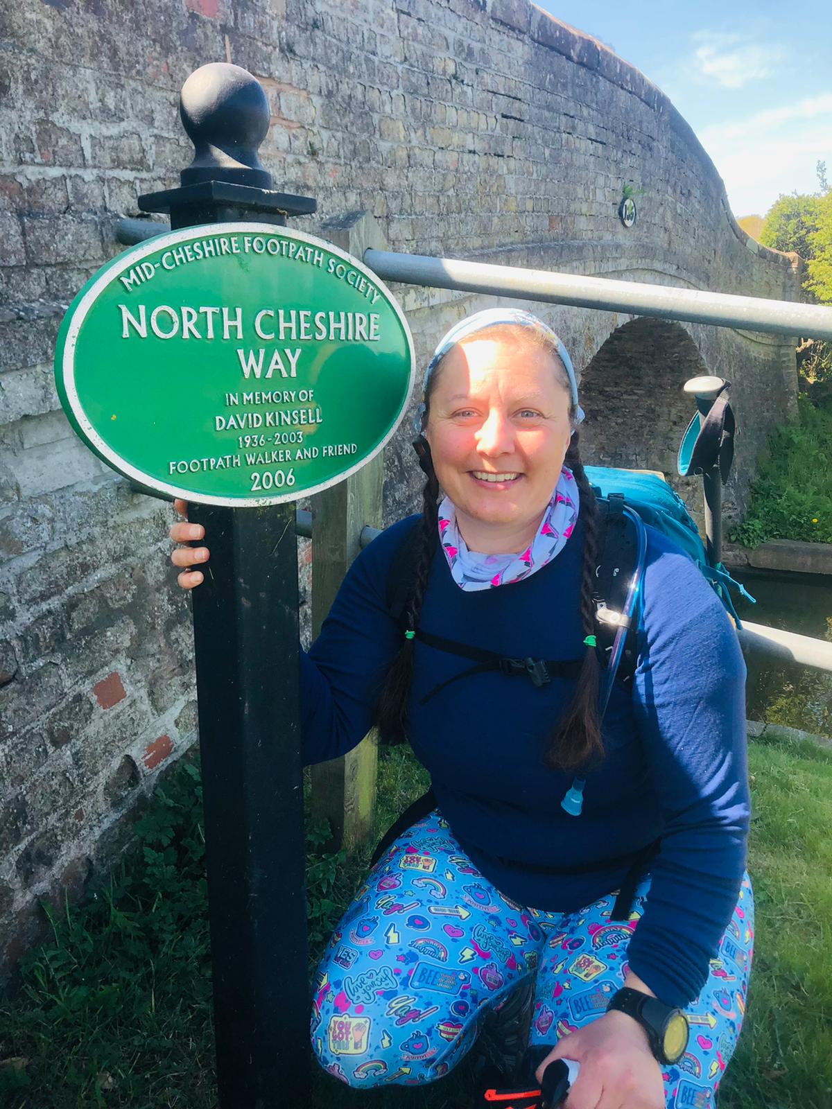 A green cast metal sign for the North Cheshire Way alongside a canal bridge. A woman in walking kit is sat next to it smiling at the camera