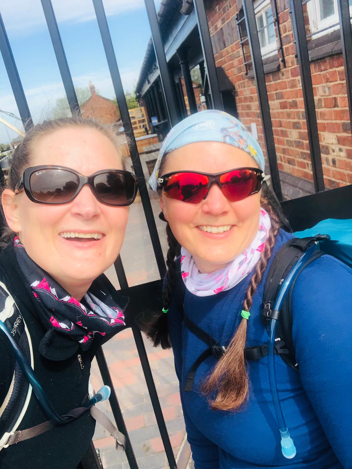 Two women in walking kit smiling in front of black metal gates