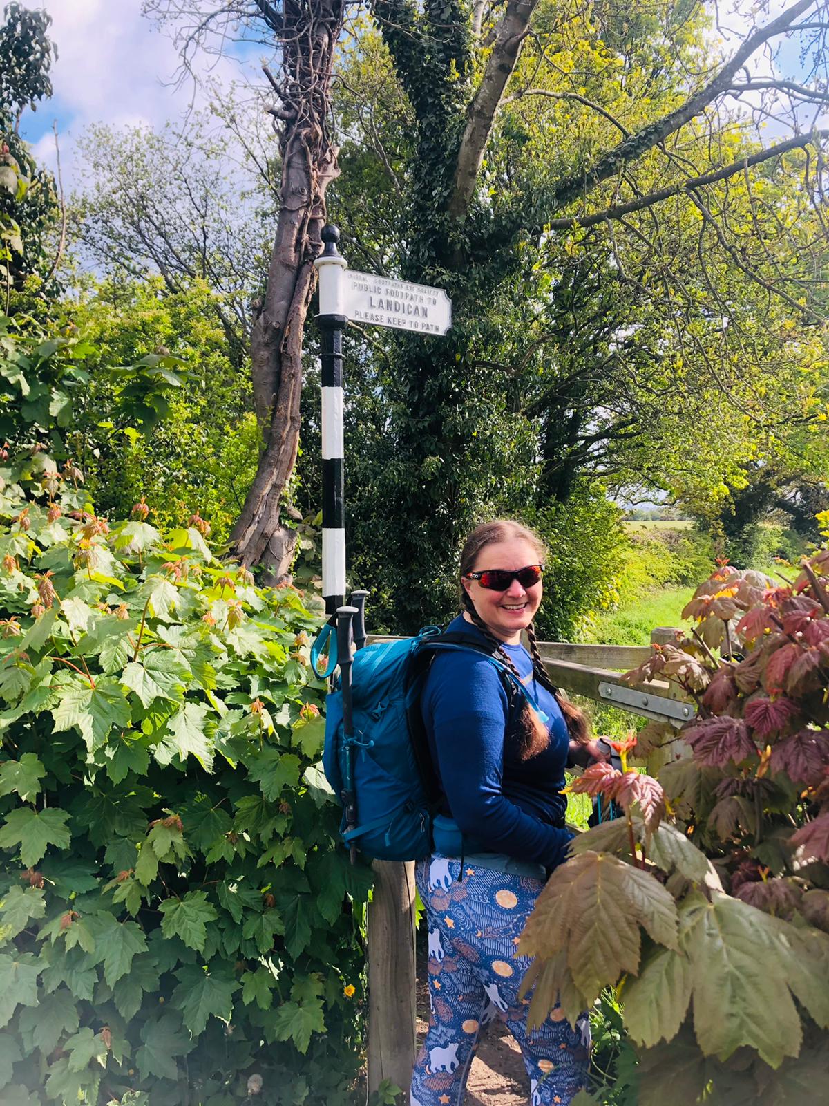 Ellen stood in front of a wooden kissing gate looks back over her shoulder to the camera. Its is sunny and the leaves are all coming out on the trees