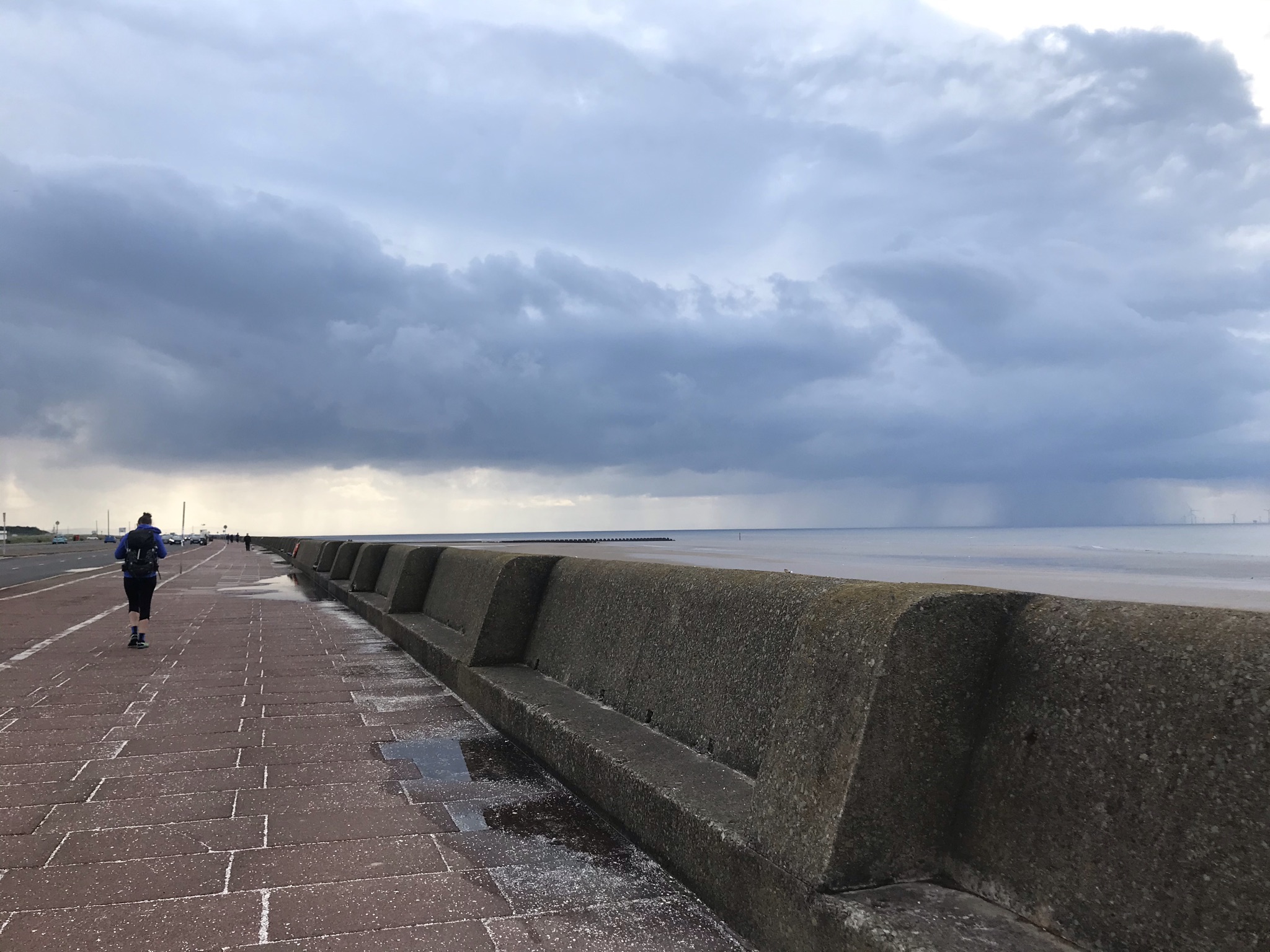 Sarah walking away from the camera along an empty promenade with a sea wall. In the far distance there is heavy rain