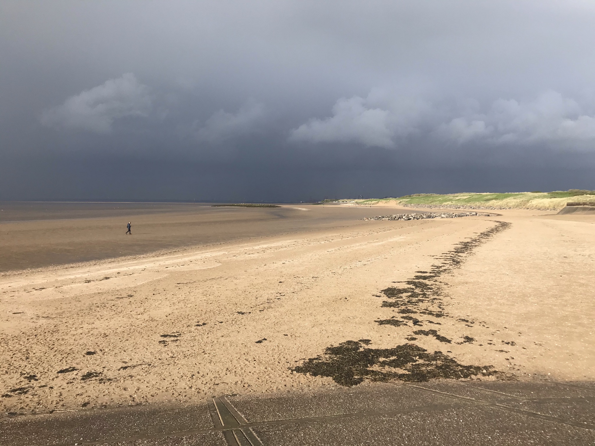 A wide sandy beach with a single person walking in the distance. The sky is dark with rain clouds