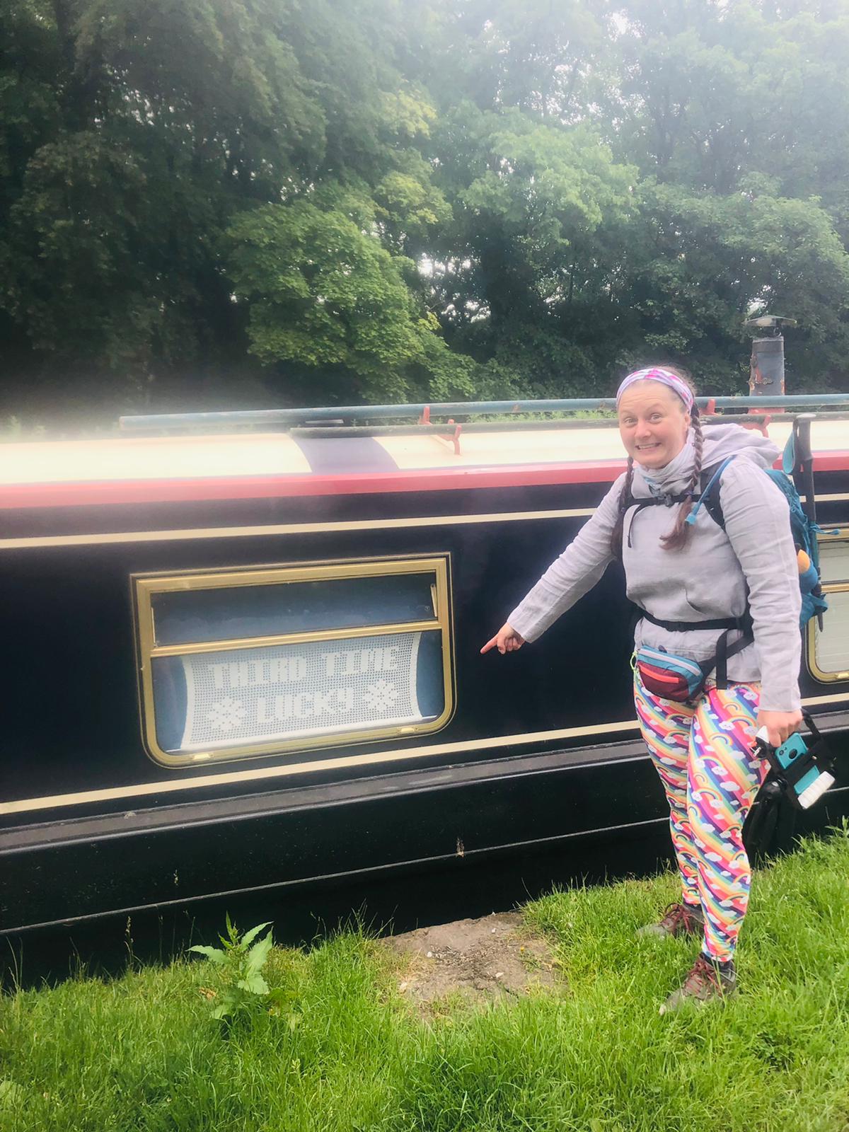 Ellen grins at the camera pointing to a lacework sign hanging in a narrowboat window that says "third time lucky"