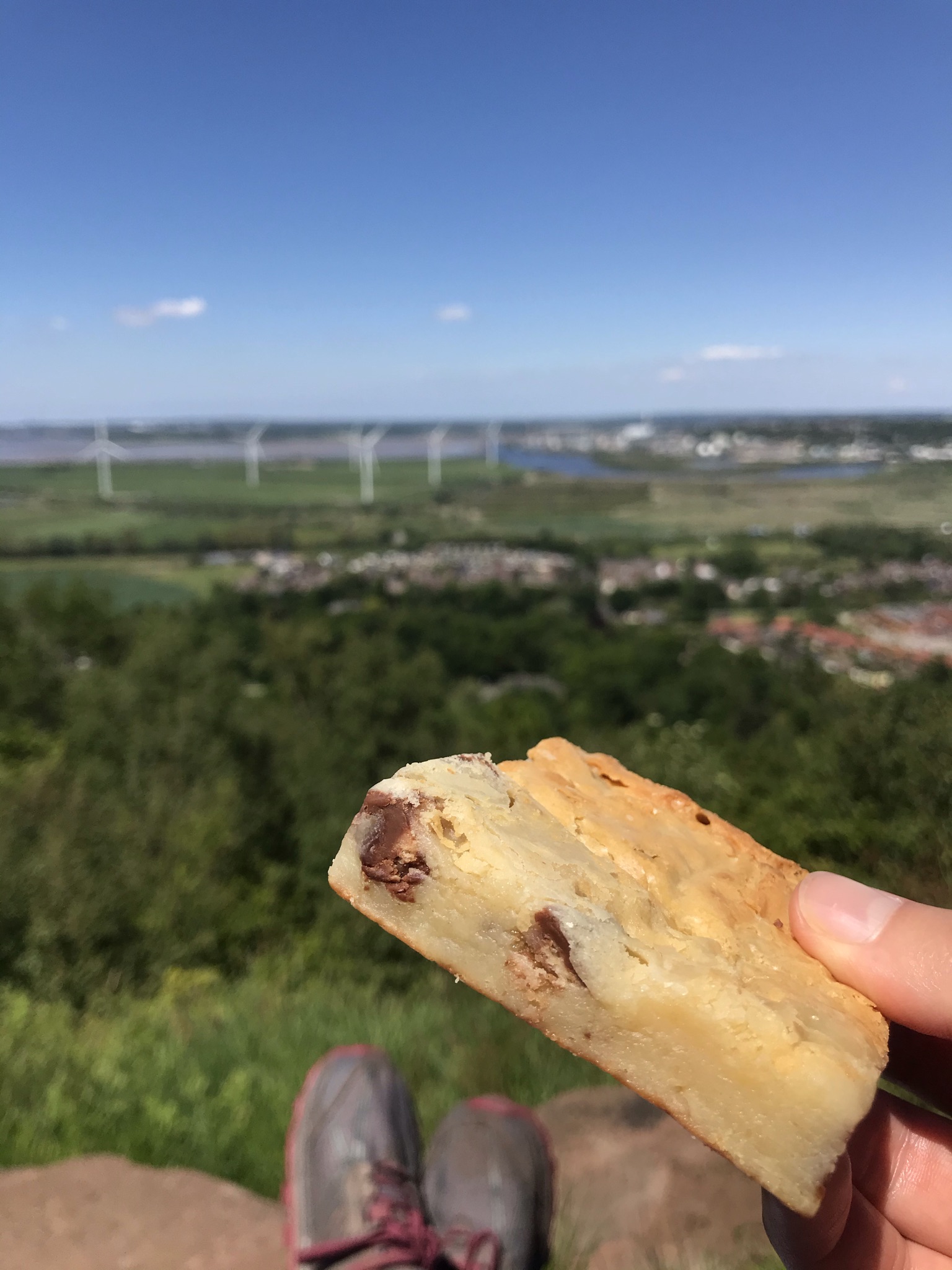 A blondie cake is held up to the camera. The background of feet resting on the stone, trees and distant wind turbines is blurred