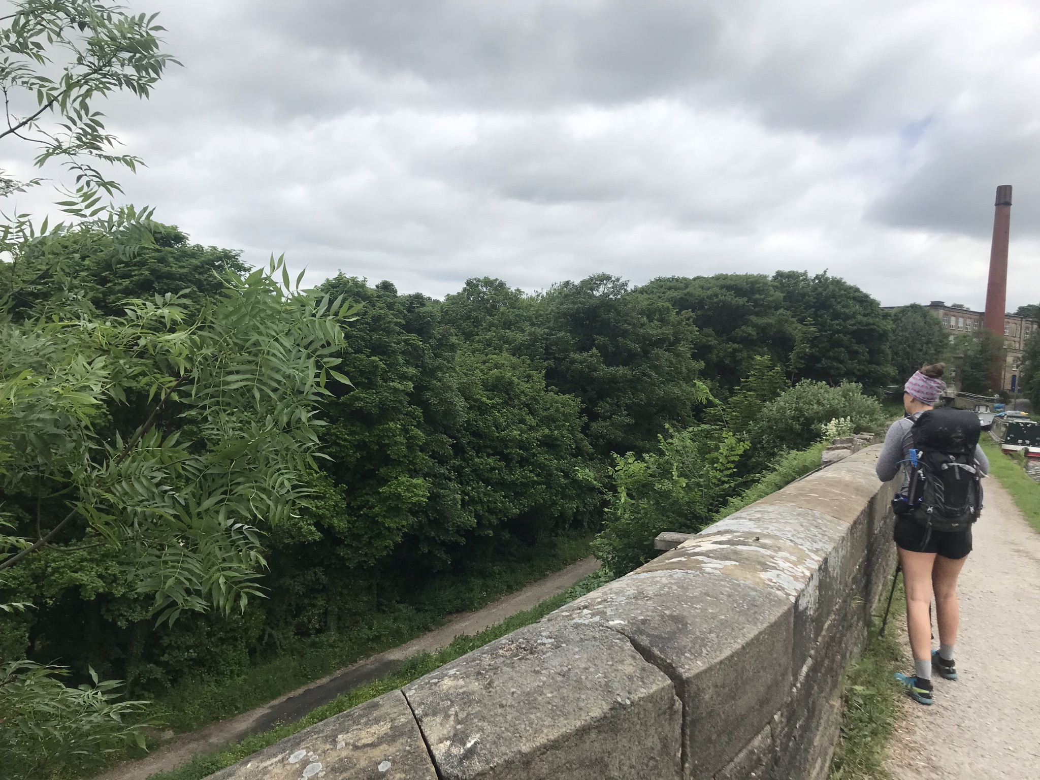 Sarah stands with her back to the camera looking over a wide stone parapet. There is a tall brick chimney and canal boats in the distance.