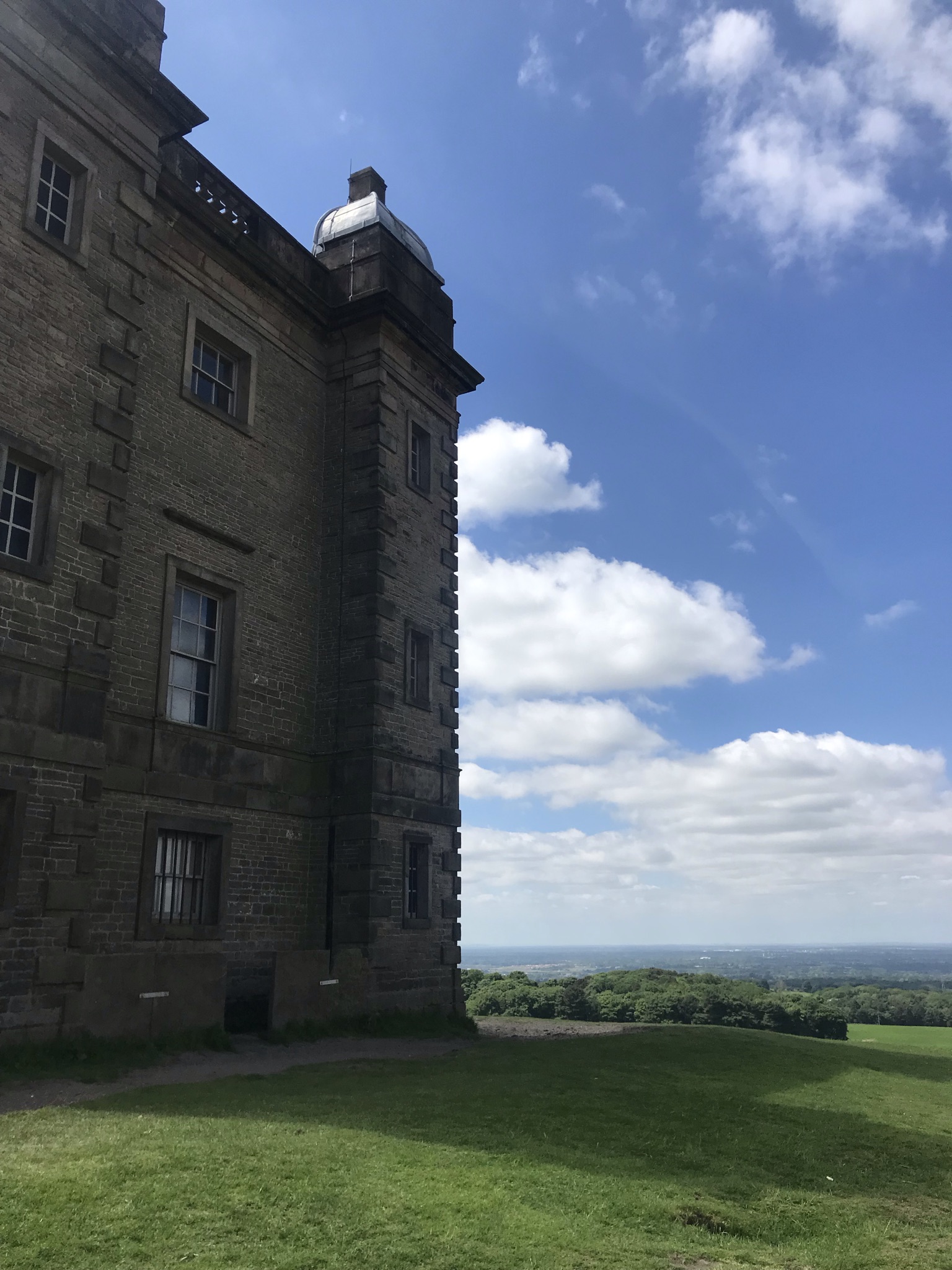 A stone 18th century three story building surrounded by short grass slopes. The sky is blue with white clouds and long views over the Cheshire plains