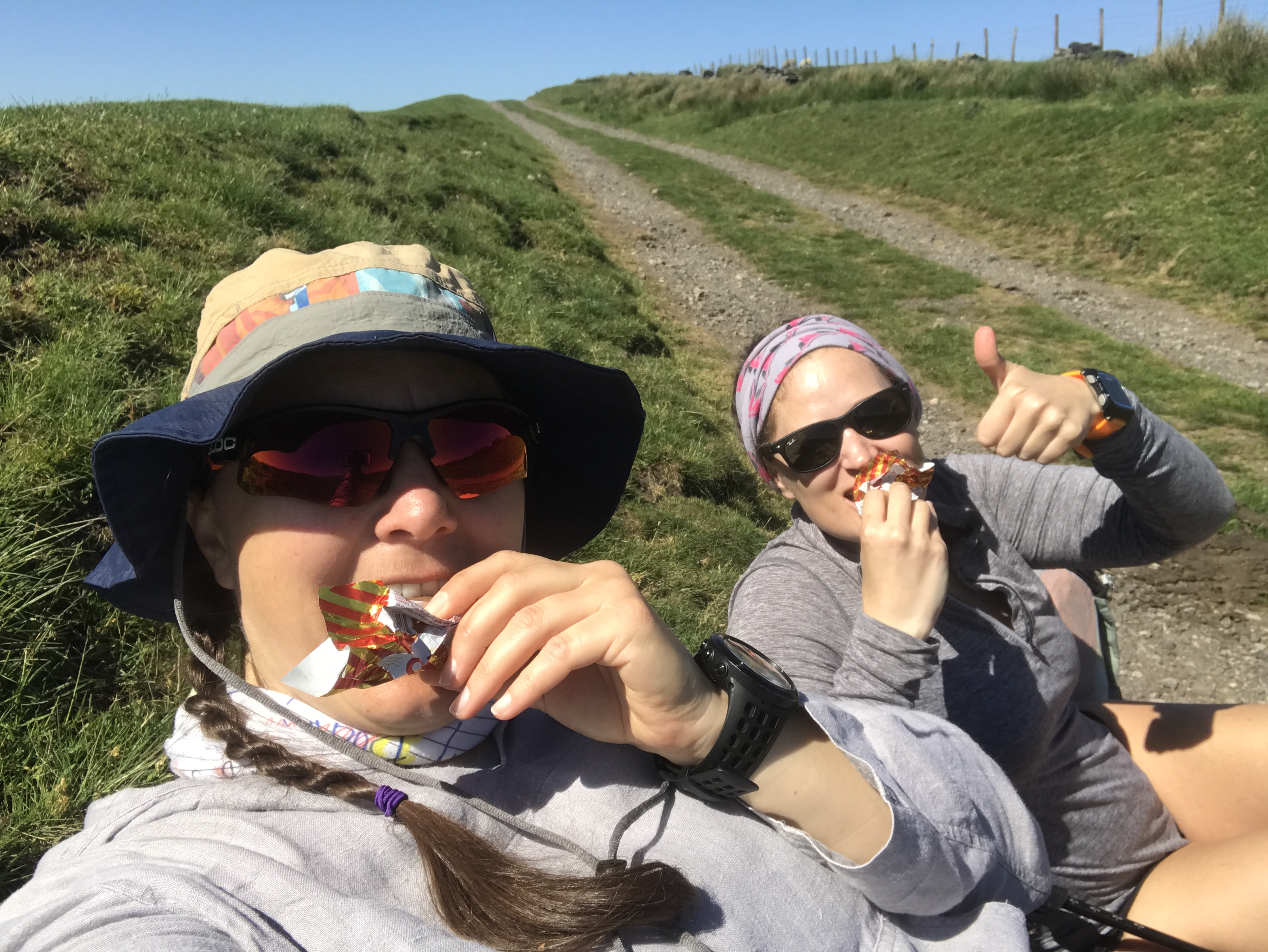 A selfie of Ellen and Sarah sat on a grass bank eating Tunnock's caramel wafer. Behind them tracks stretch up to a blue sky