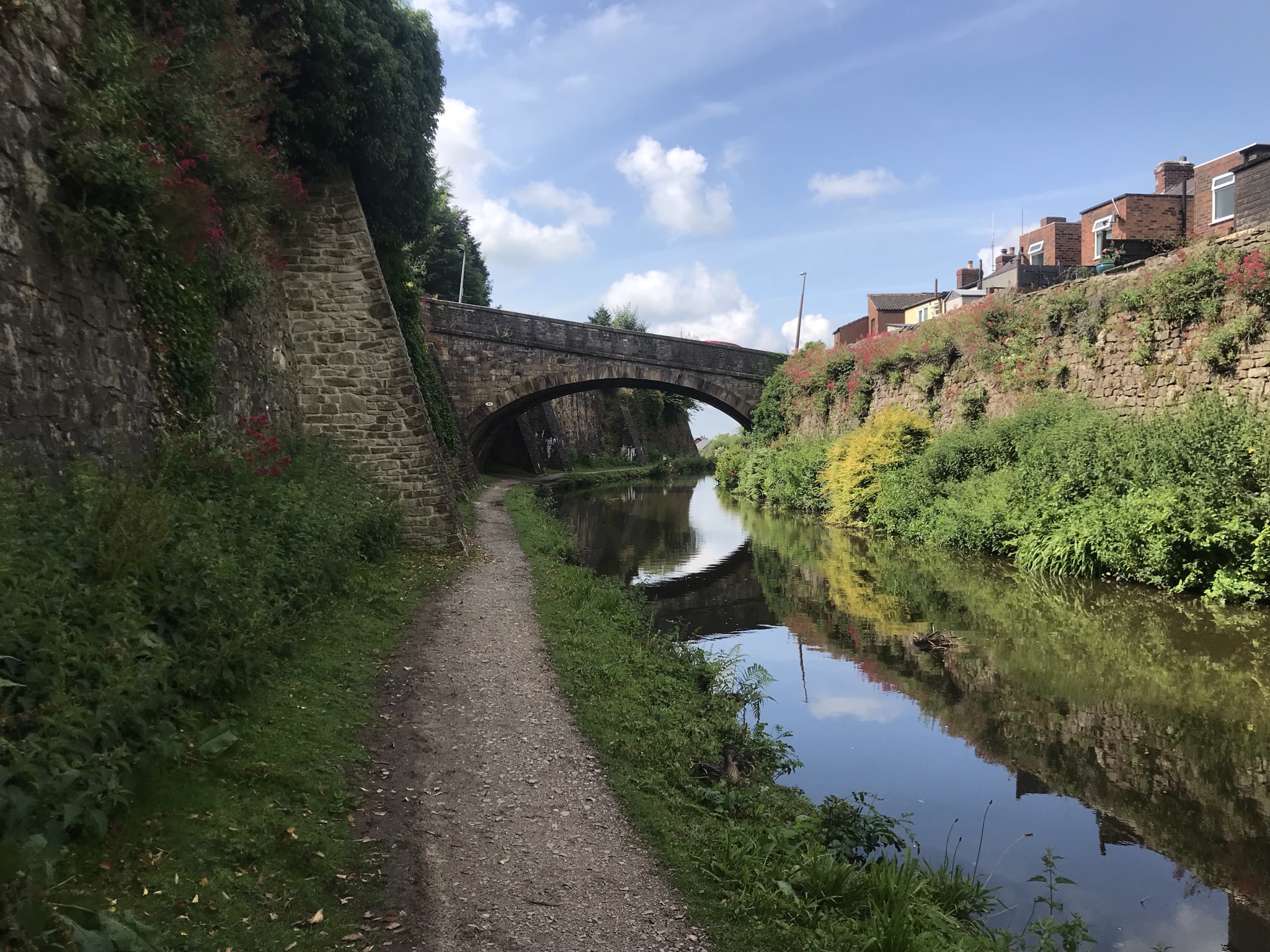 A towpath along the canal with high, flower covered stone walls either side. The canal is still and reflecting the walls and the sky