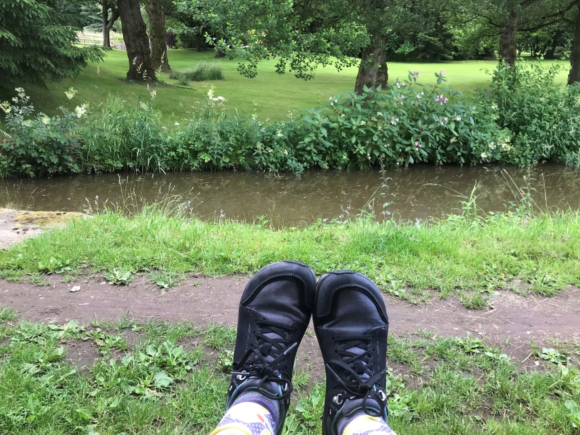 Feet with grey lace up walking shoes held up with the canal and neat mown grass on the far bank