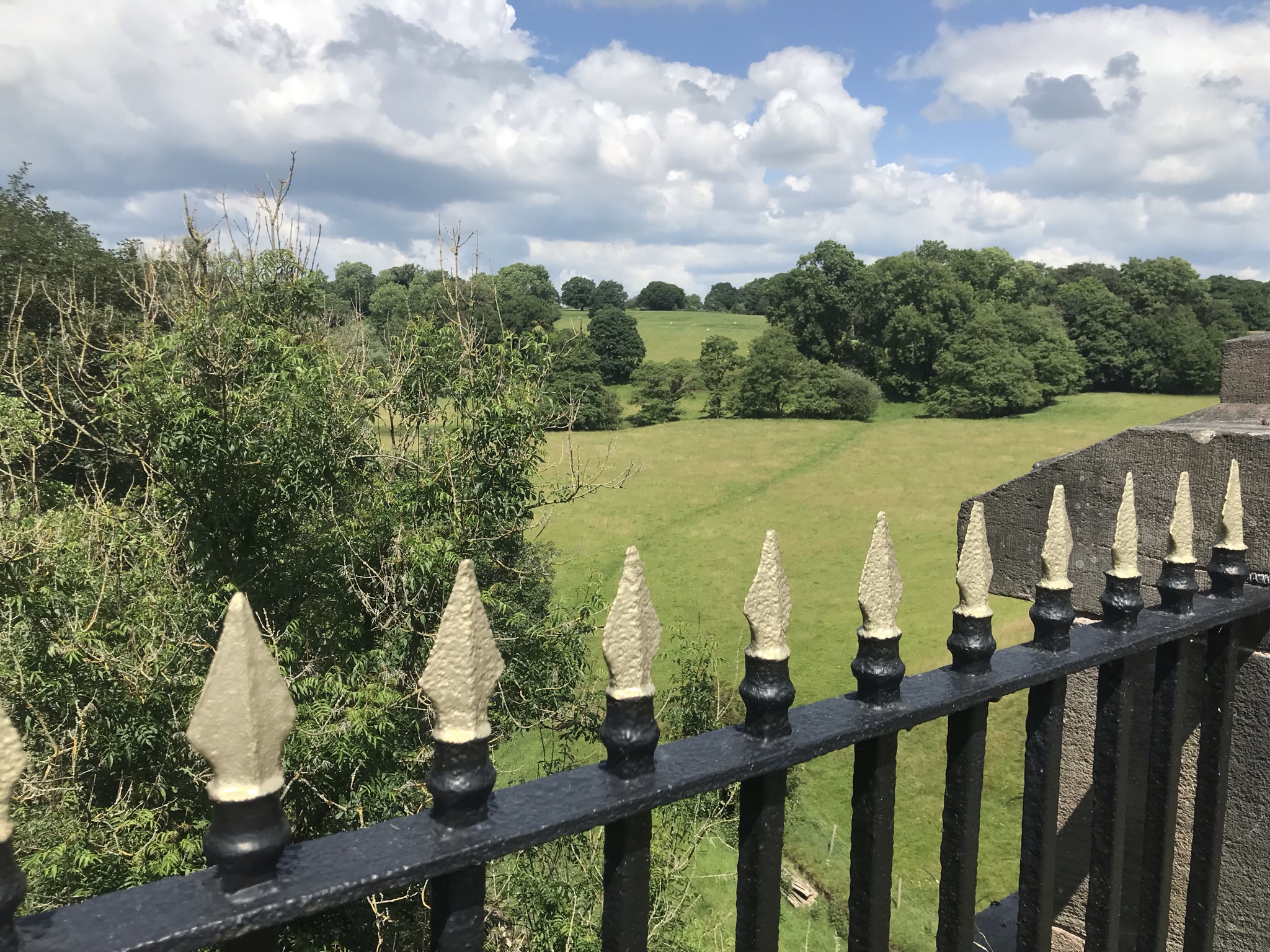 Gold and black railings overlooking a long drop to green fields and trees, with fluffy clouds in the sky