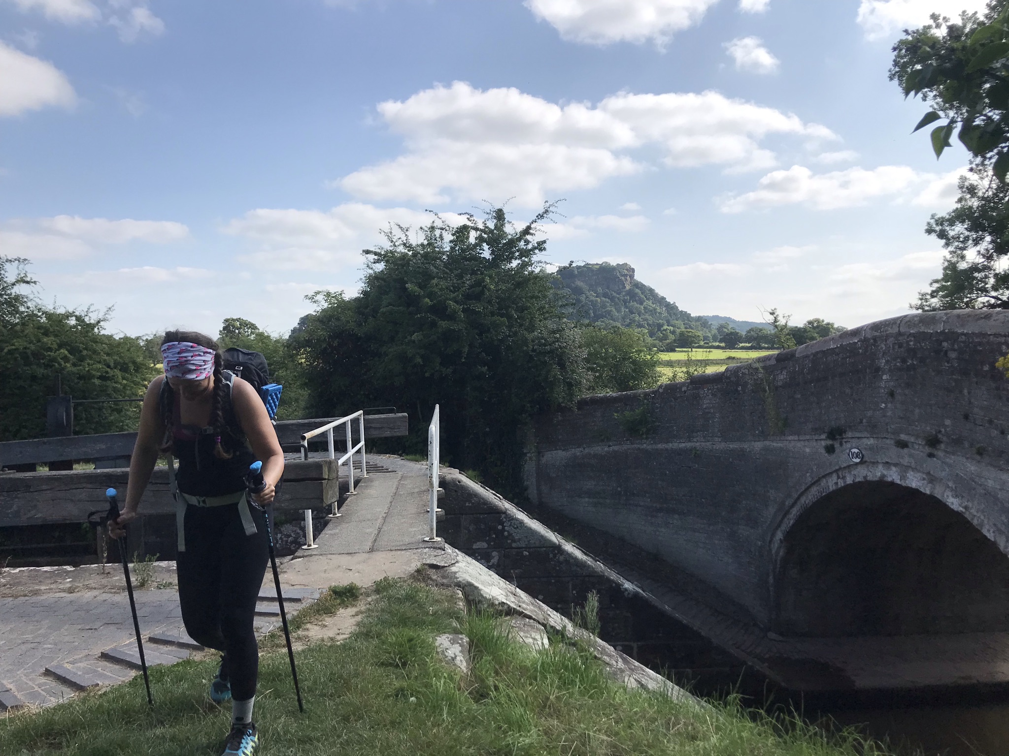 Sarah, walking with poles crosses a narrow bridge over a lock. Beeston Castle is in the distance