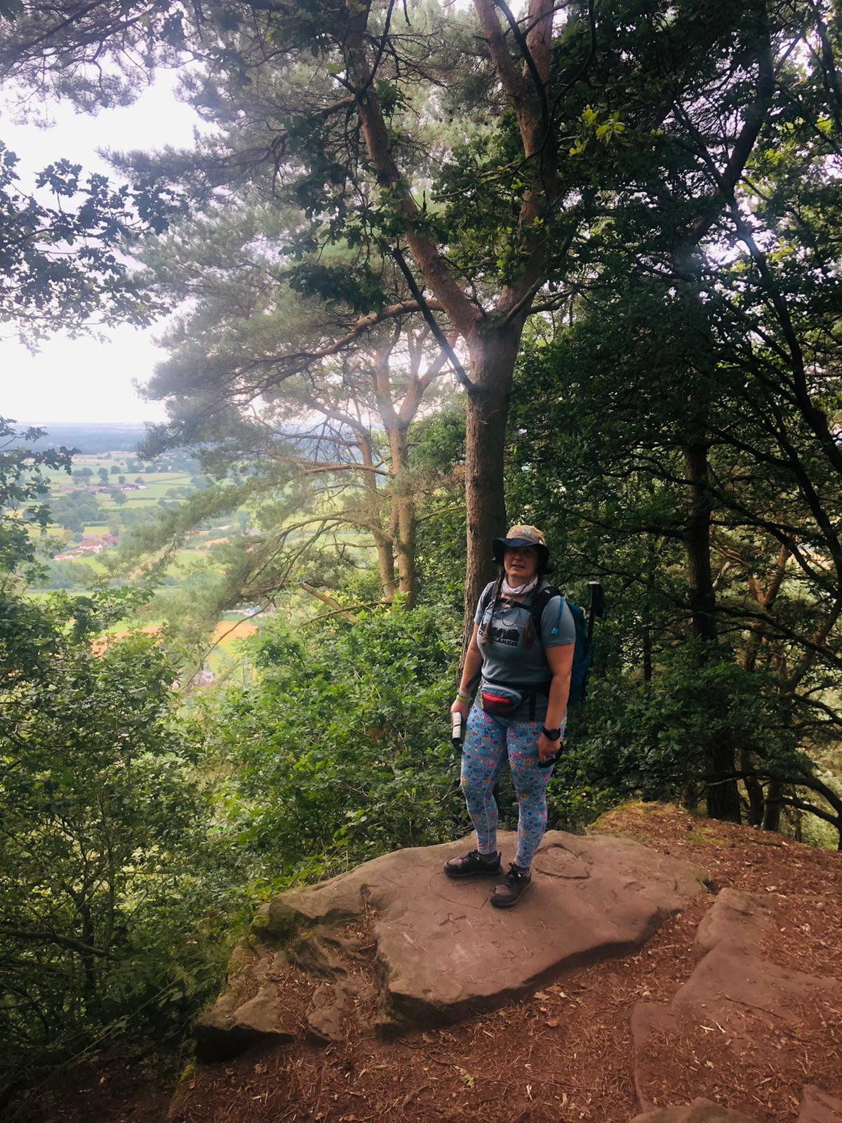 Ellen stands on a rock outcrop surrounded by trees. There is a long view across fields below.