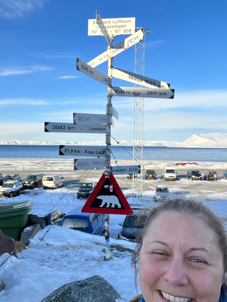 A selfie in front of the signpost showing the distance to major cities in the world.  At the bottom, there is the famous polar bear warning sign