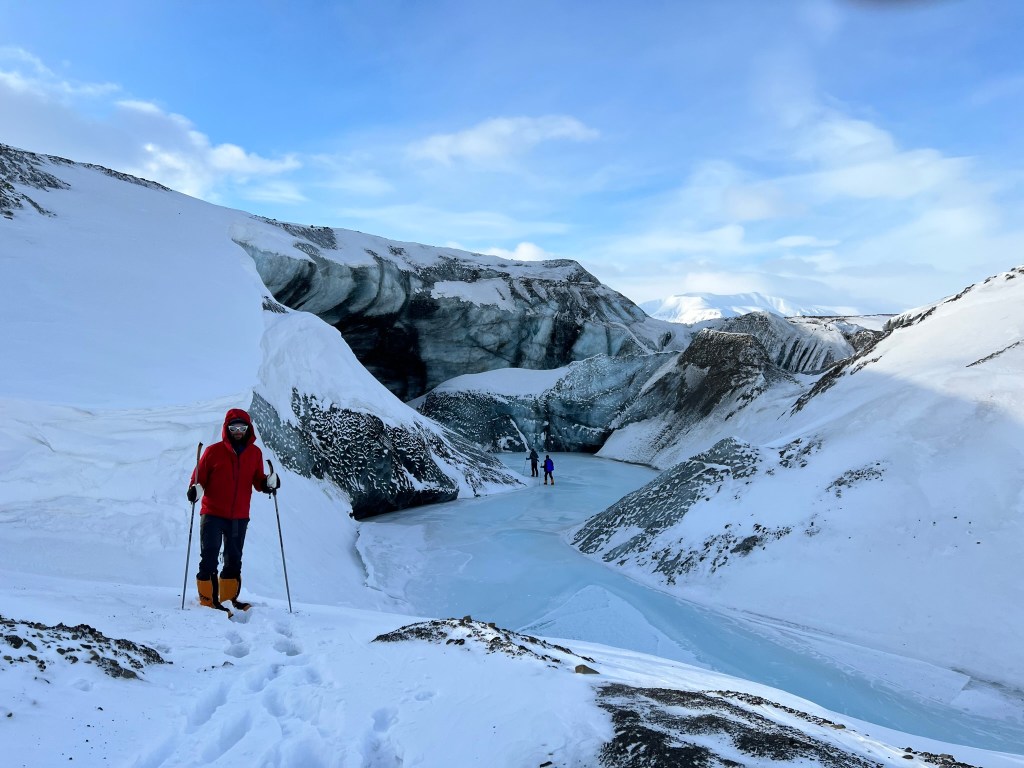 A skier in a red jacket stands above the the steep drop to the frozen river flowing out from under the glacier wall