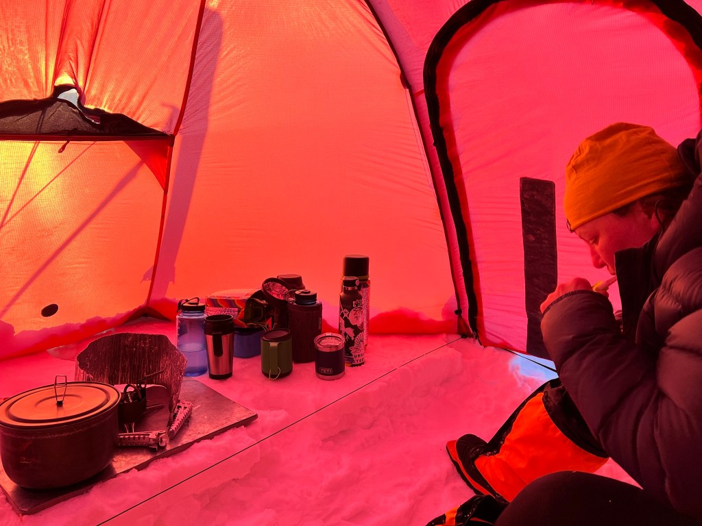 Me sat in my down jacket and arctic boots, cleaning my teeth. Opposite me is the petrol stove and pots, and a collection of mugs and flasks.