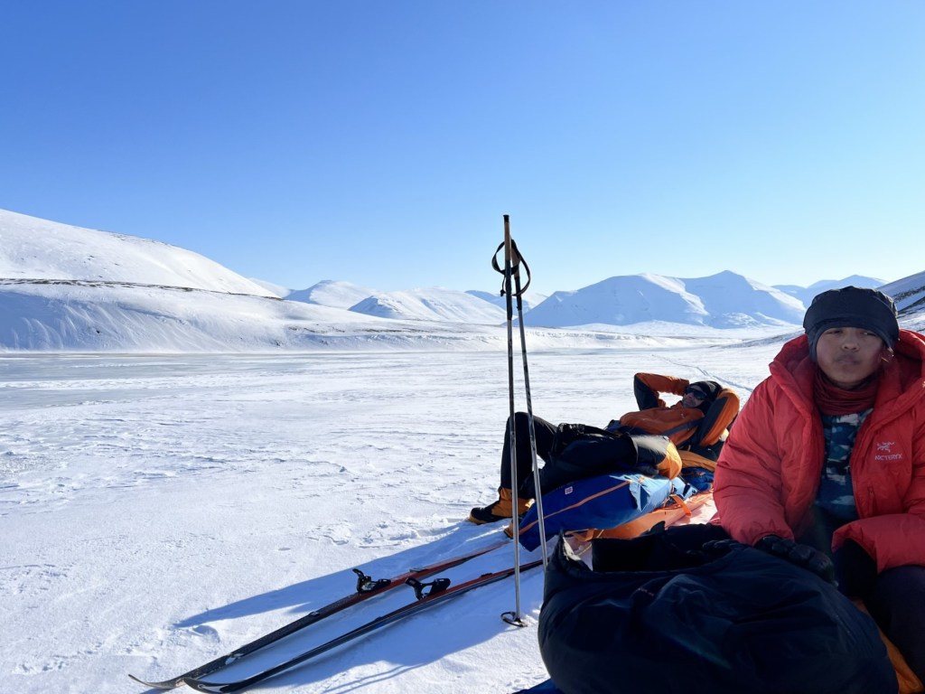 Team members relaxing on their pulks in the sun, with our track stretching off in the distance down the valley